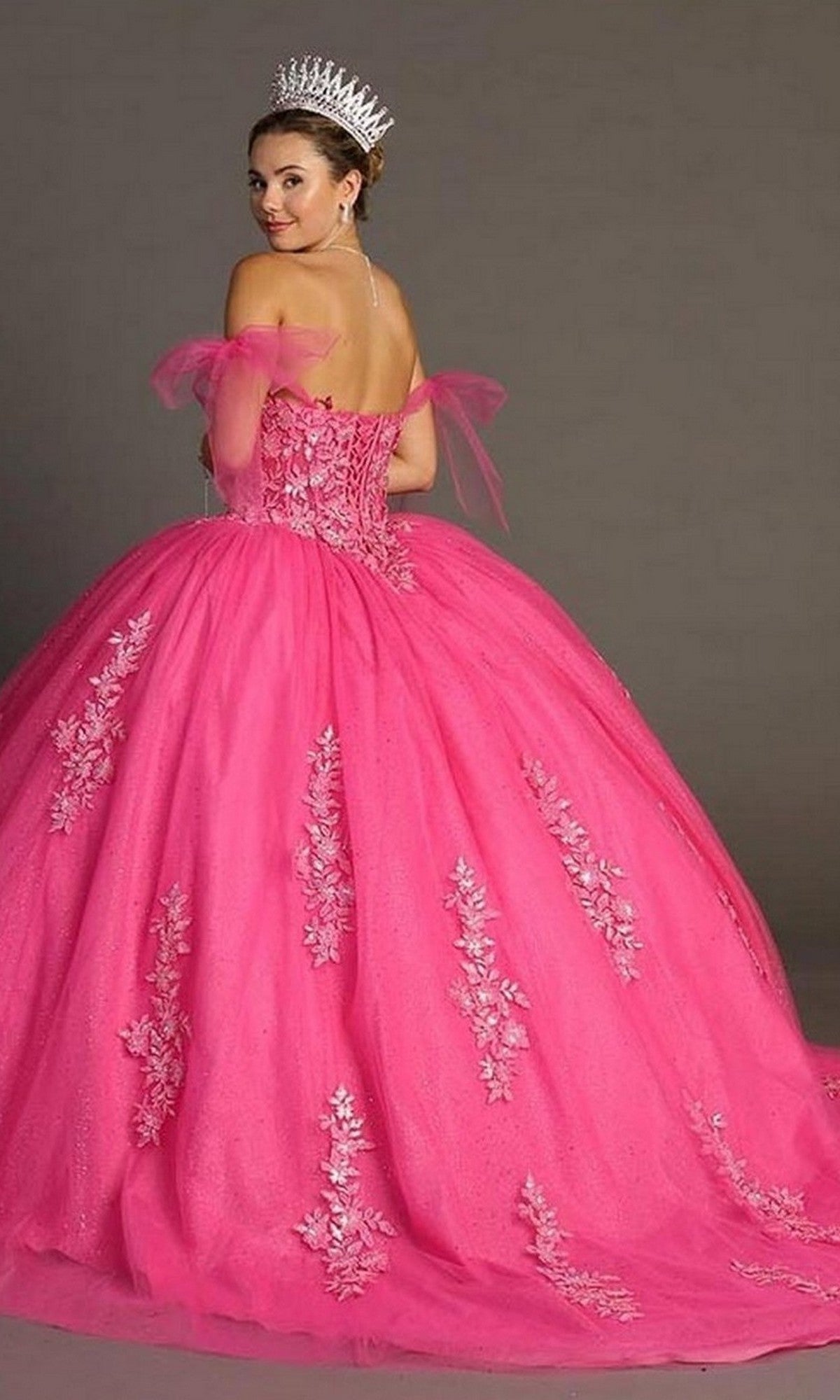 A woman wearing the Quinceanera Dress CHF3086 by Chicas—a bright pink off-the-shoulder ballgown skirt adorned with lace details and a tiara—stands against a neutral background, glancing over her shoulder.